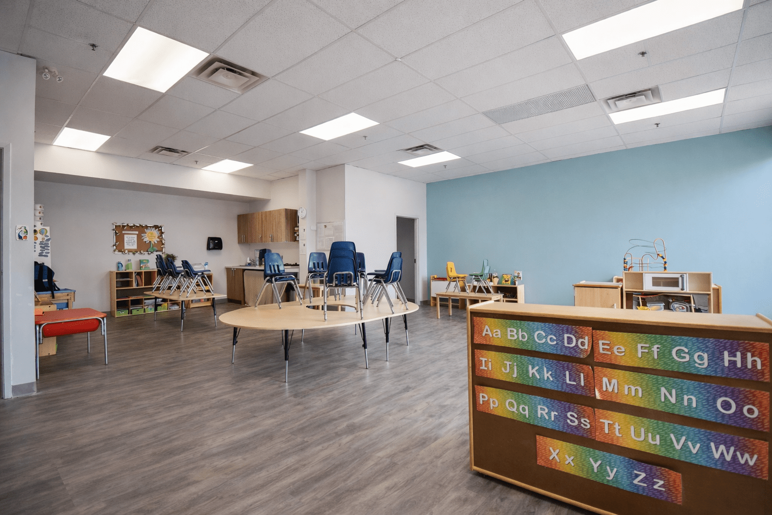 Open-concept daycare classroom with child-height furniture, commercial vinyl flooring, acoustic ceiling system, and integrated kitchenette for early childhood education space