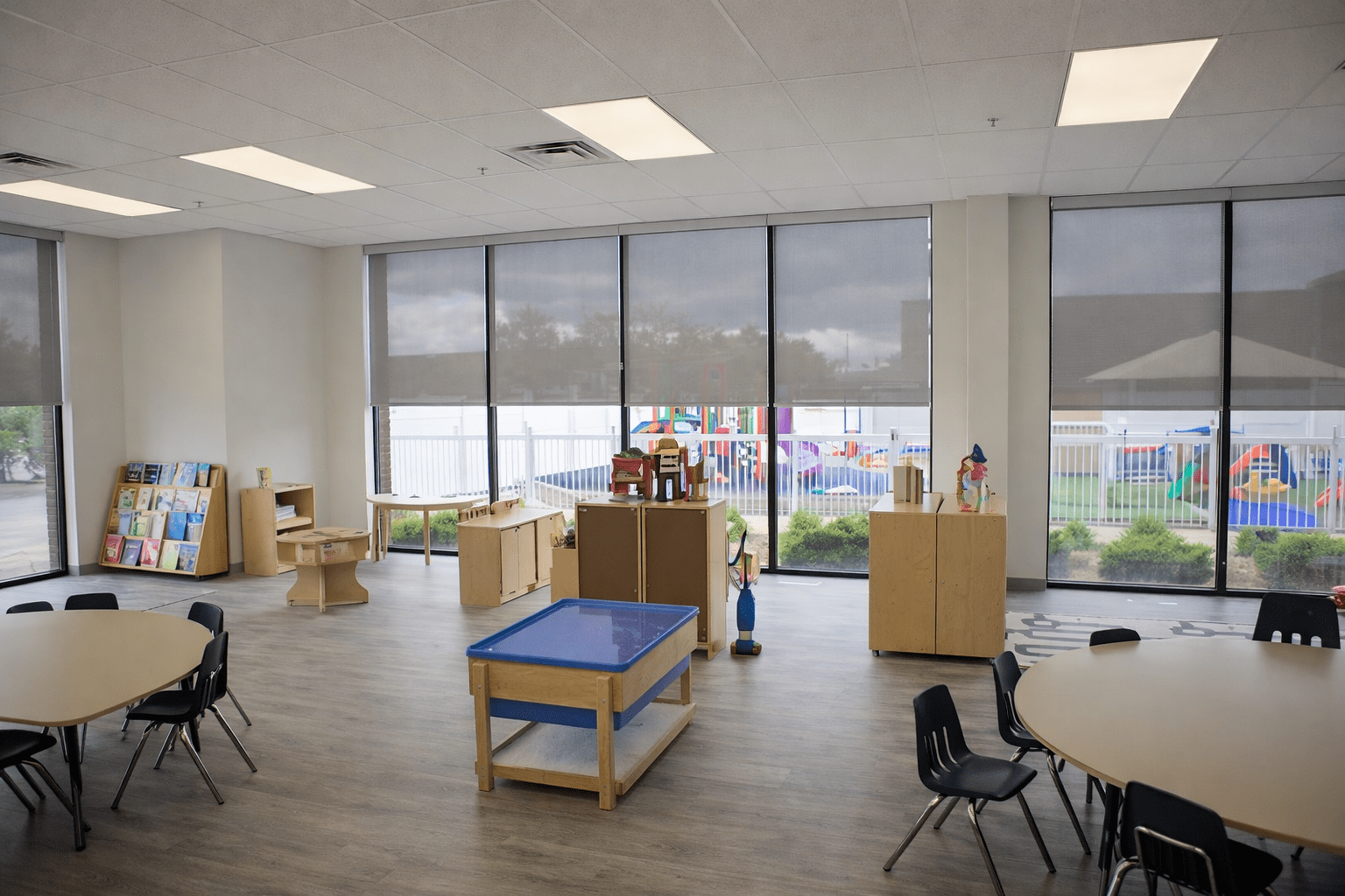 Modern daycare classroom interior with floor-to-ceiling glazing, natural light, vinyl plank flooring, and child-safe millwork storage units in a licensed childcare facility