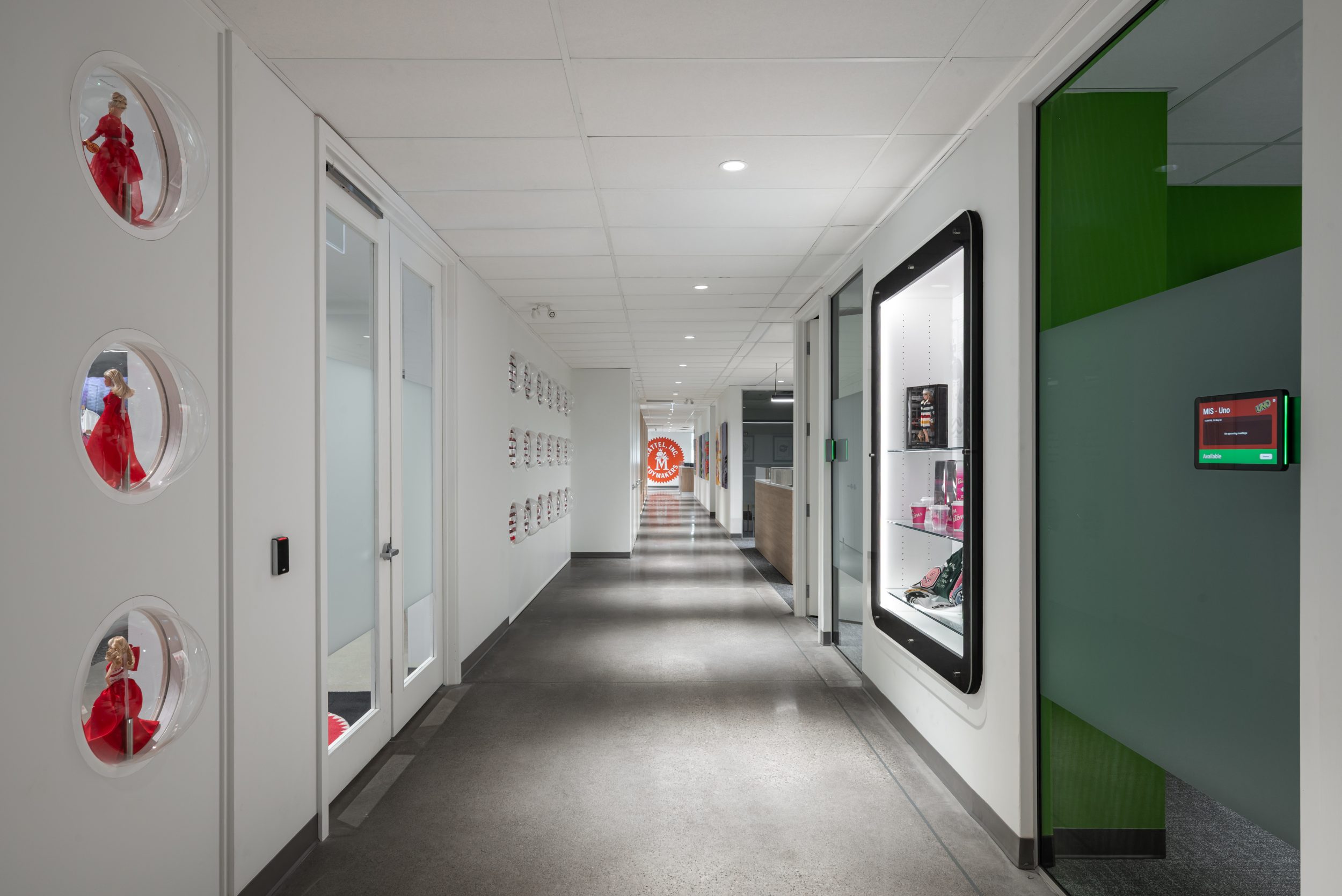 Branded corporate office corridor at Mattel Canada Headquarters in Mississauga, Ontario, featuring custom product display cases, glass-front meeting rooms, polished concrete floors, architectural lighting, integrated room-booking technology, branded interior graphics, and contemporary commercial office construction.