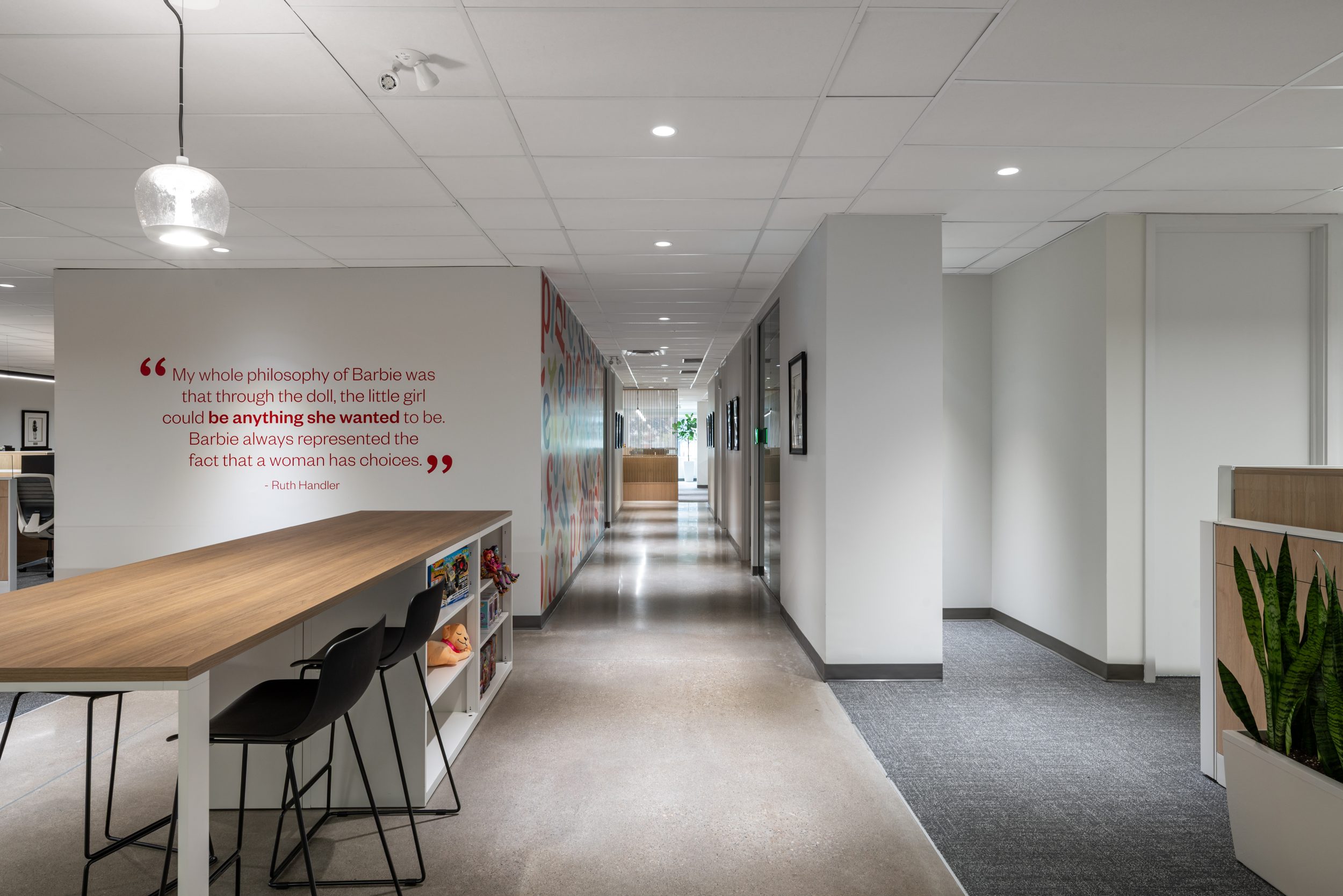 Collaborative touchdown workspace and modern office corridor at Mattel Canada Headquarters in Mississauga, Ontario, featuring custom millwork, built-in shelving, bar-height seating, polished concrete flooring, branded wall graphics, architectural lighting, and contemporary commercial office renovation completed through a Design-Build delivery model.